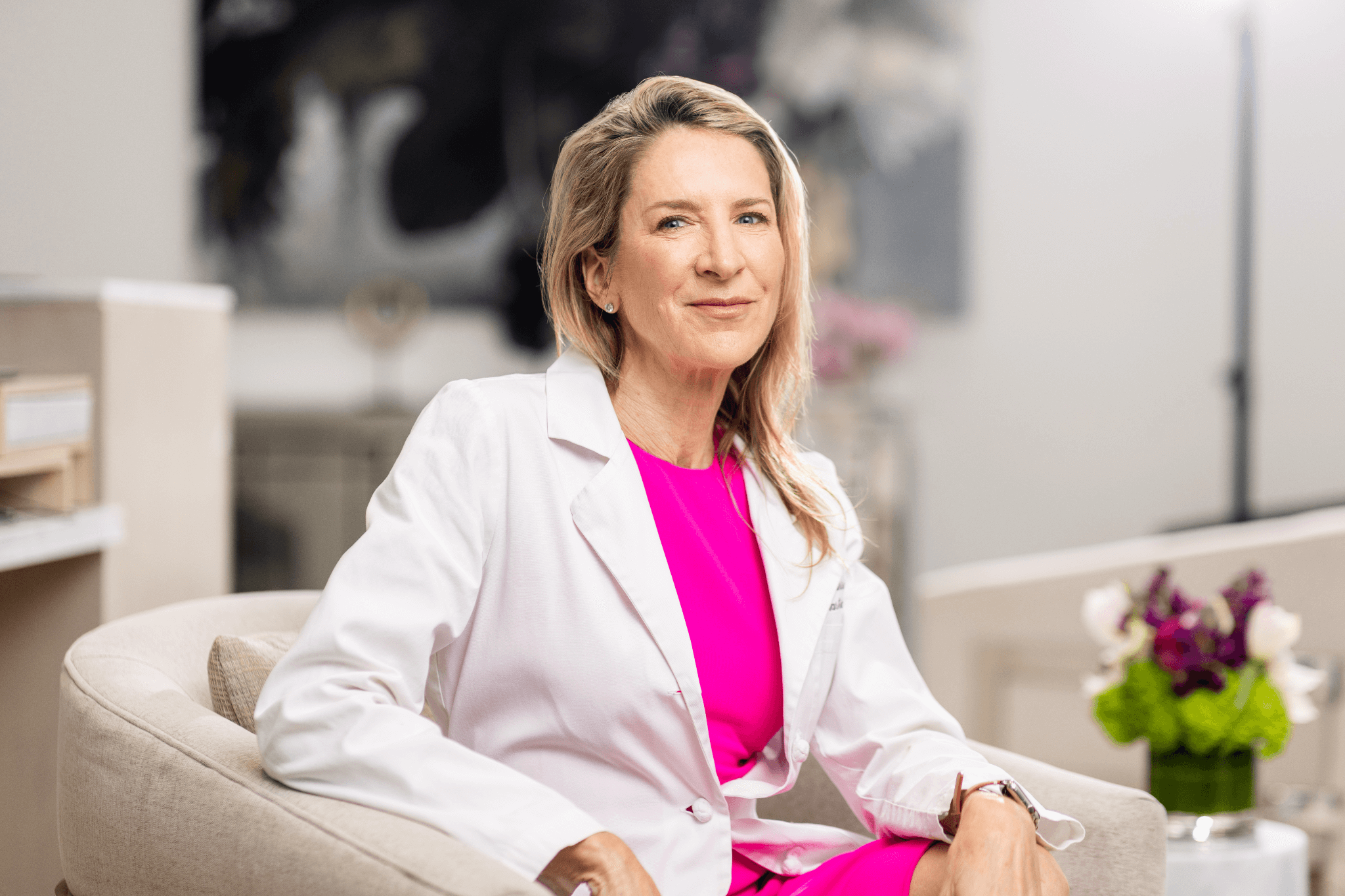 Dr. Lisa Cassileth in a white coat and bright pink dress, seated in a modern clinic setting and smiling confidently